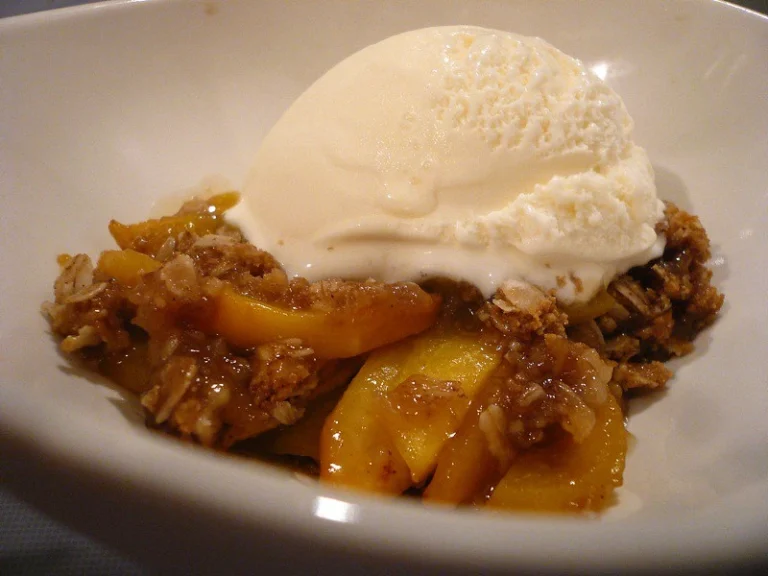 A golden brown peach crumble served in a baking dish, topped with a buttery crumble and garnished with a scoop of vanilla ice cream.