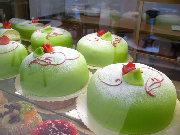 A beautifully decorated Swedish princess cake with a smooth green marzipan covering, dusted with powdered sugar, topped with a pink marzipan rose and leaf, sitting on a white plate.