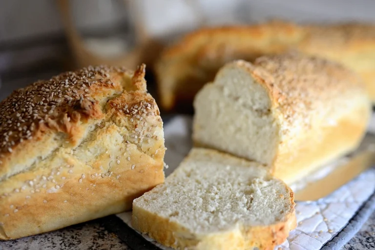 A freshly baked round loaf of sheepherder bread with a golden-brown crust, sitting on a wooden board surrounded by a few wheat stalks.