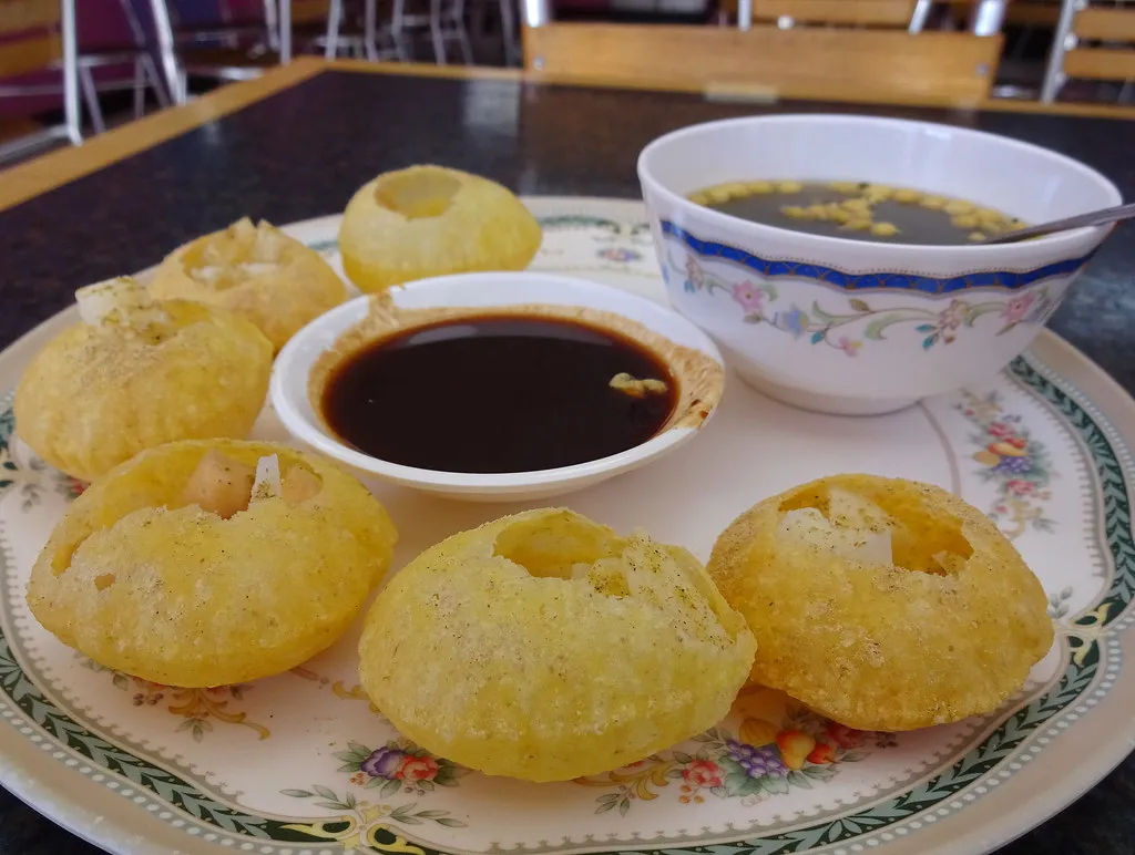 A plate of crispy, hollow puris filled with spicy, tangy tamarind water, mashed potatoes, and chickpeas, served with a side of mint-flavored pani in a traditional Indian street food style.