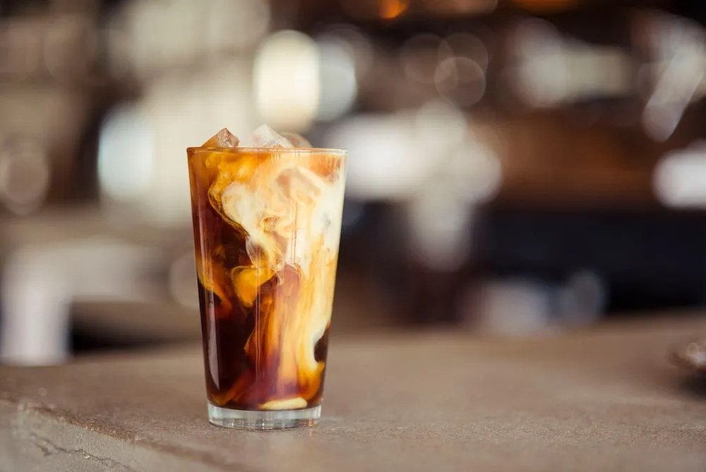 A refreshing iced latte in a tall glass with ice cubes, creamy swirls of milk mixing with espresso, and a straw, set on a wooden table.