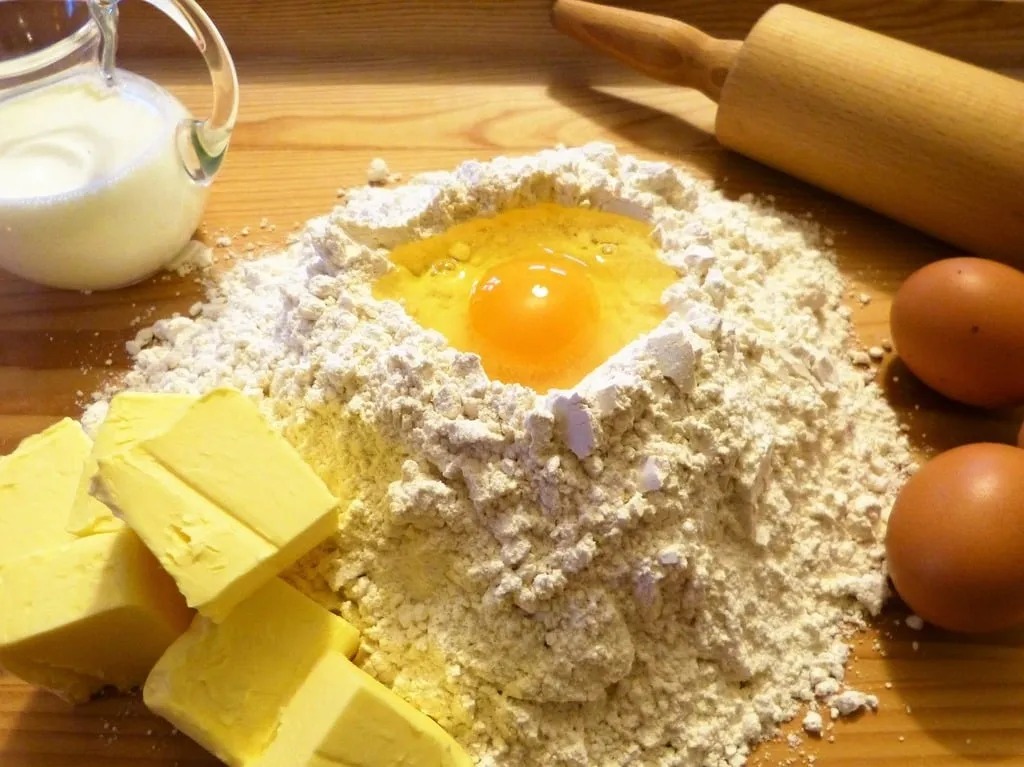 Baking essentials for coffee cake: a bowl of flour, a stick of butter, a glass of milk, eggs, and a small dish of sugar.