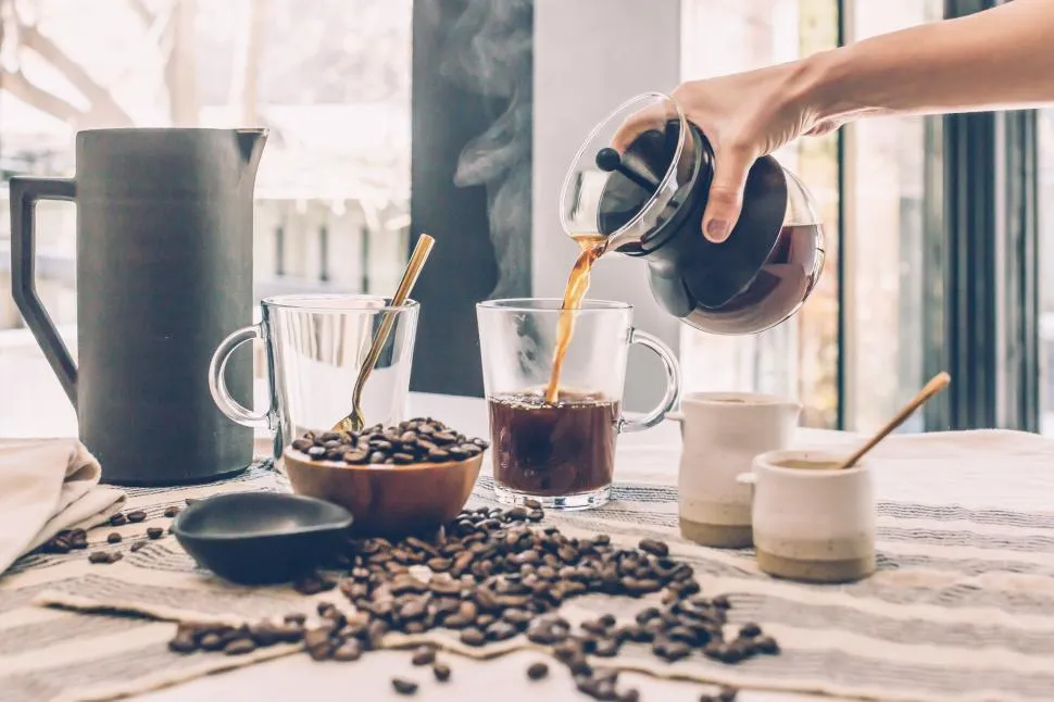 A steaming cup of freshly brewed French press coffee in a glass mug, placed on a wooden table with coffee beans scattered around.