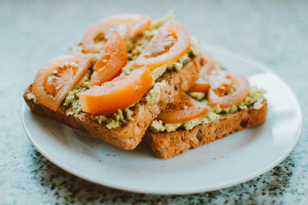 Toasted sourdough bread topped with mashed avocado, tomato slices, and sprinkled herbs on a white plate.