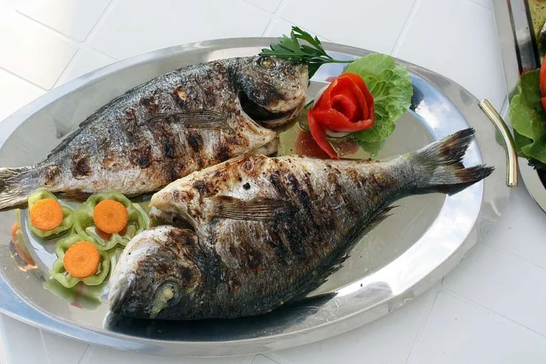 Freshly cooked fish fillet with lemon slices, herbs, and seasoning on a plate, served with vegetables.