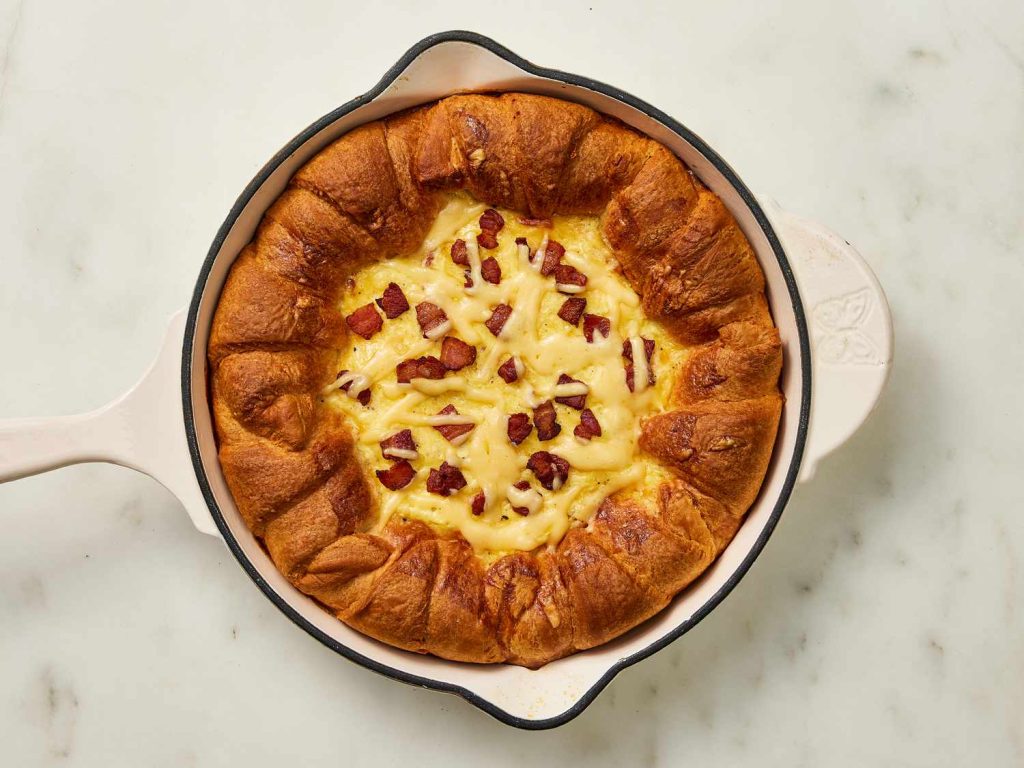 Golden, flaky croissant breakfast pie filled with crispy bacon and melted cheese, served on a rustic wooden plate.