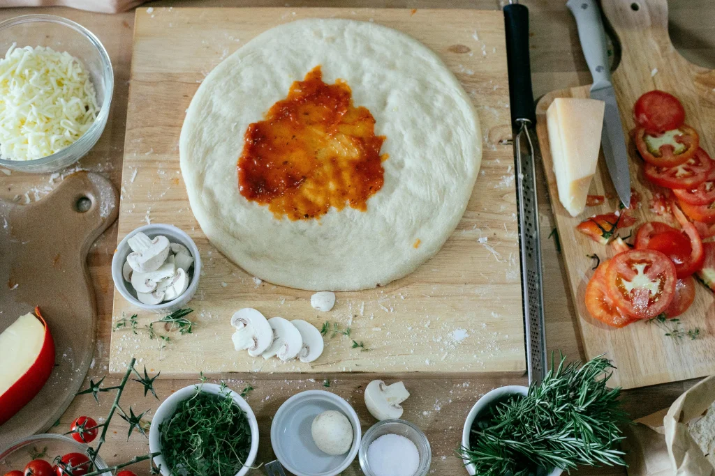 "Close-up of focaccia pizza dough being topped with fresh ingredients like cheese, herbs, and tomatoes before baking