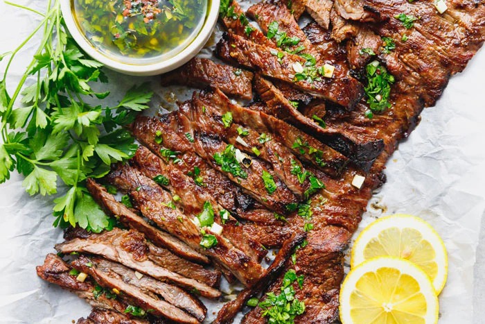 Grilled skirt steak with a smoky char, sliced and served on a rustic wooden board with fresh herbs and a side of sauce.