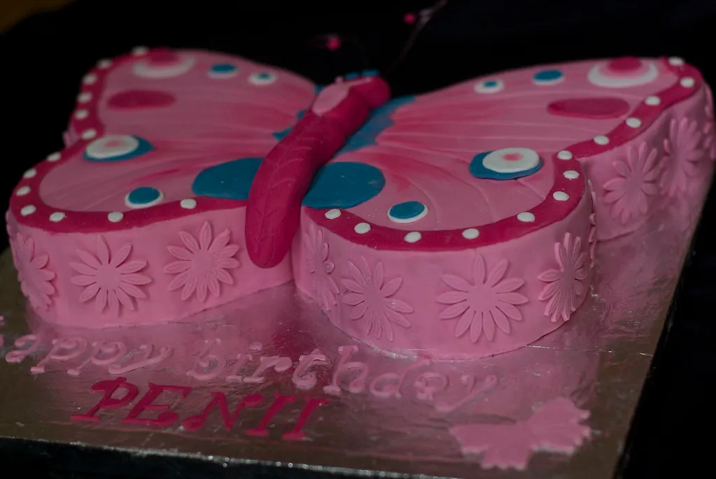 Whimsical butterfly cake decorated with vibrant, pastel-colored wings made of buttercream, adorned with delicate sugar flowers and edible glitter, set against a soft, elegant backdrop
