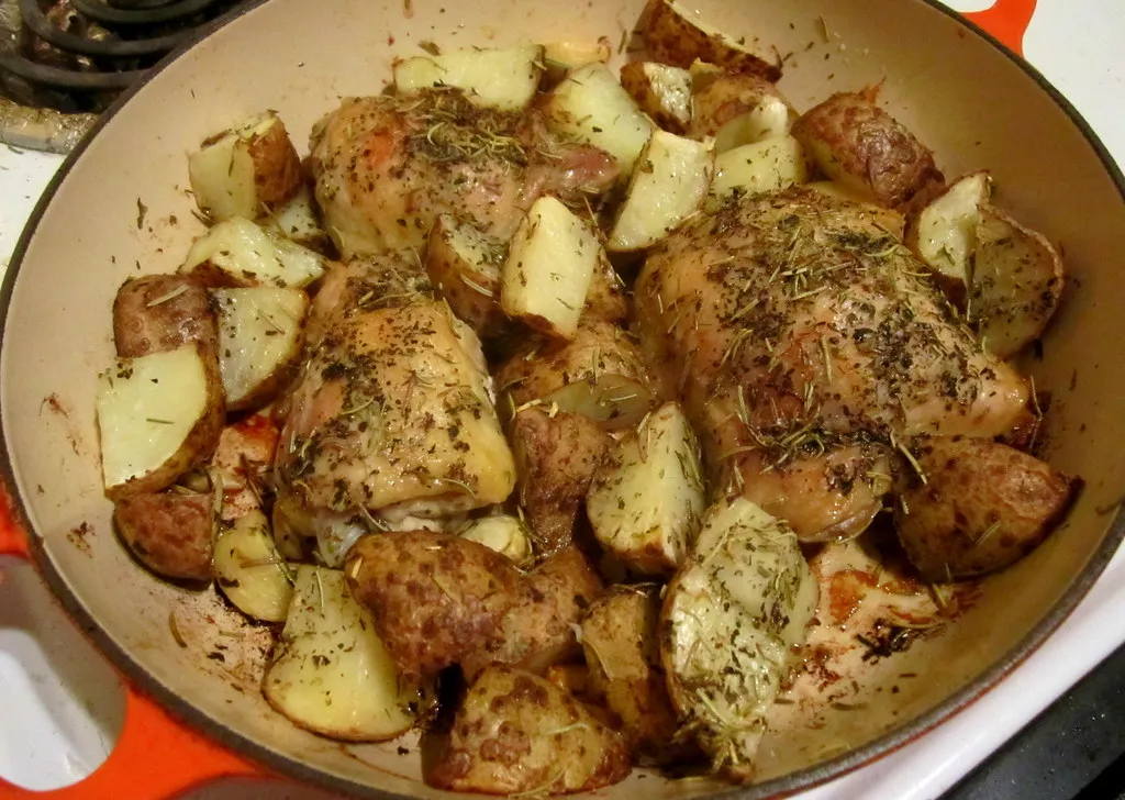 Golden roasted Greek lemon chicken thighs with crispy skin served alongside herb-seasoned roasted potato wedges on a rustic white plate