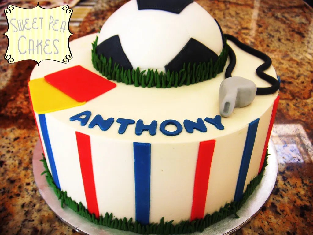 Soccer cake decorated with a realistic soccer ball design, complete with green grass icing, a white and black pattern resembling a soccer ball, and small details like a goalpost and players.