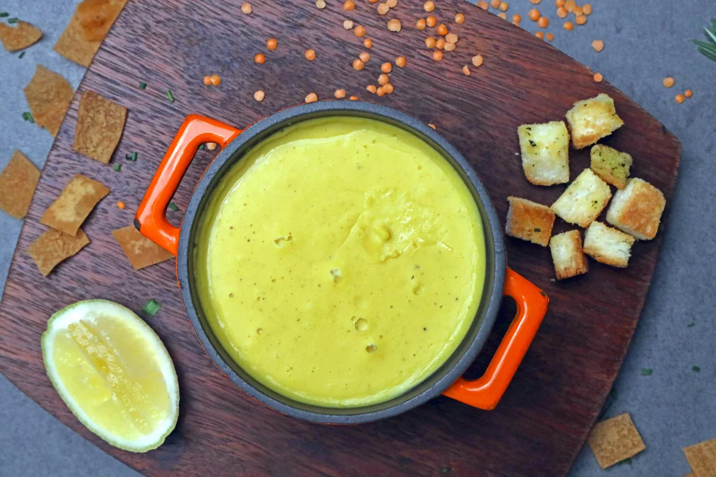 Flat lay of raw soup ingredients including carrots, onions, celery, garlic, spices, and a pot of broth on a rustic wooden surface.
