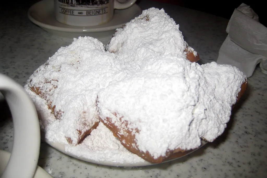 "Freshly fried French Quarter-style beignets dusted generously with powdered sugar, served hot on a plate with a cup of chicory coffee