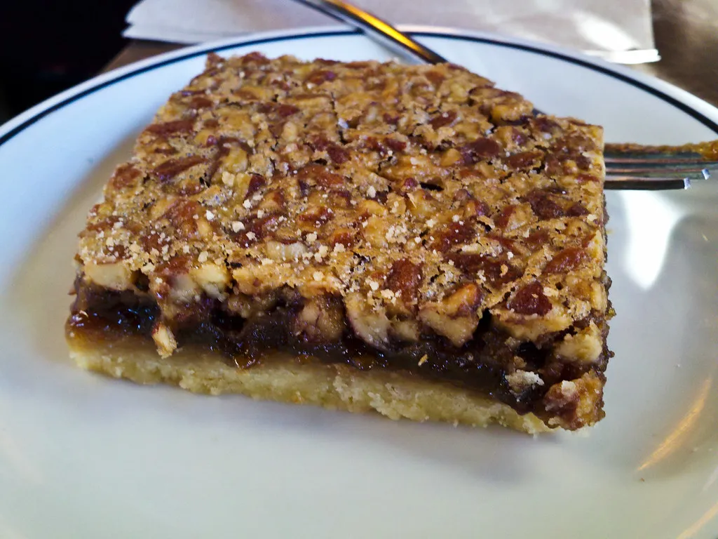 Chewy pecan pie bars with a golden crust, topped with chopped pecans and a glossy spicy glaze, served in neat squares on a rustic tray
