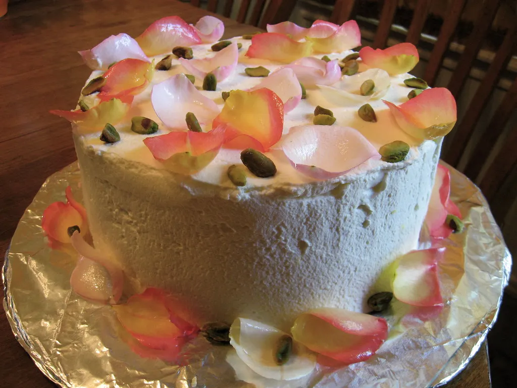 A slice of Persian Love Cake decorated with crushed pistachios and rose petals, showcasing its moist texture and golden color infused with saffron and cardamom.