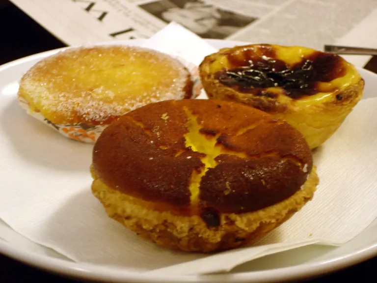 Assorted Portuguese breakfast pastries including Pastéis de Nata, Pão de Deus, and Bola de Berlim arranged on a wooden board with coffee cups in the background.