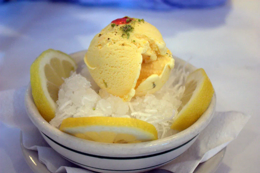 A colorful serving of Persian ice cream topped with pistachios and rose petals next to a bowl of Faloodeh Shirazi, a traditional Iranian frozen dessert made of thin vermicelli noodles mixed with rosewater syrup and lime, both presented on a rustic wooden table.