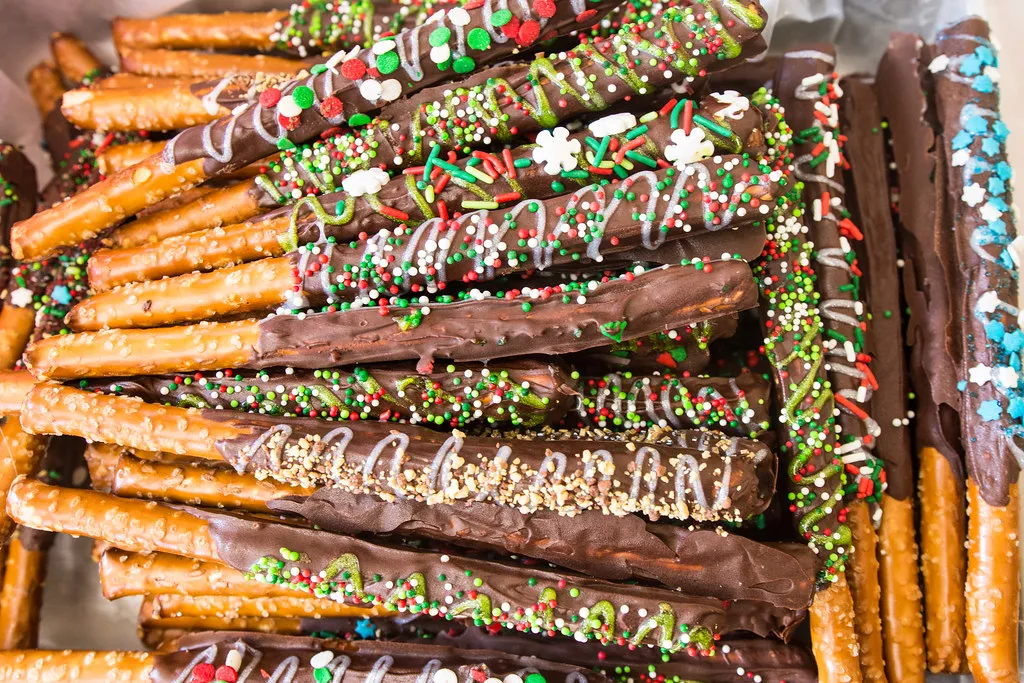 Colorful candy-dipped pretzel rods coated with sprinkles, neatly arranged in a glass jar on a party dessert table.