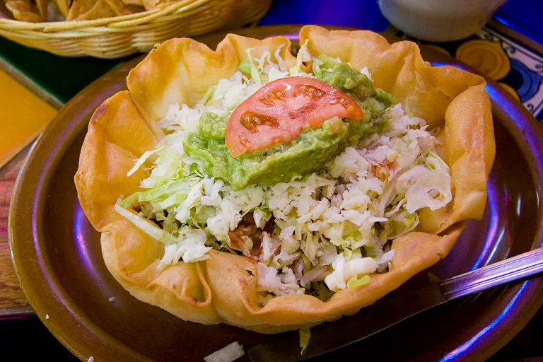 Traditional taco salad with ground beef, lettuce, tomatoes, cheese, sour cream, and tortilla chips in a white bowl.