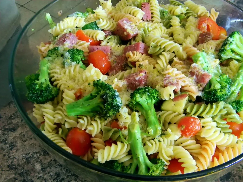 A glass container filled with colorful cold pasta salad made with rotini, cherry tomatoes, cucumbers, and herbs, placed on a wooden table beside a serving spoon.