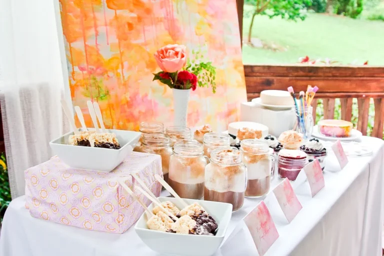 Elegant dessert table setup with assorted mini cakes, macarons, cupcakes, and floral decor in a pastel and gold theme.