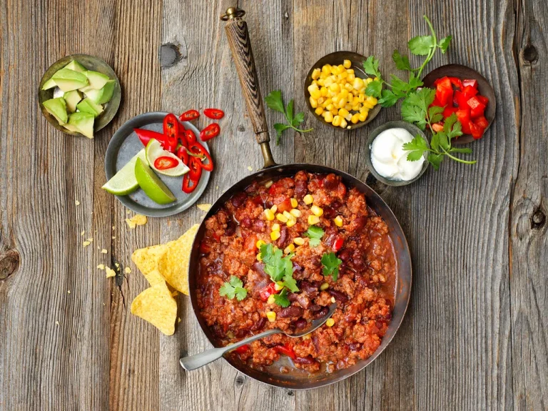 A colorful spread of spicy Mexican dishes including chili con carne, salsa roja, salsa verde, tacos, and jalapeños, all served on a rustic wooden table.