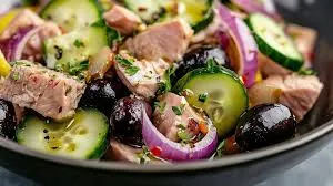 Colorful Mediterranean tuna salad with chickpeas, cherry tomatoes, cucumbers, olives, and feta served in a white bowl with lemon wedges and parsley on the side