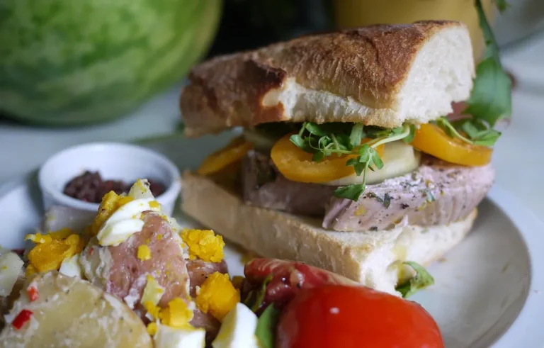 Close-up of a perfectly made tuna sandwich with lettuce and tomato on toasted whole grain bread, served on a wooden board with chips on the side.
