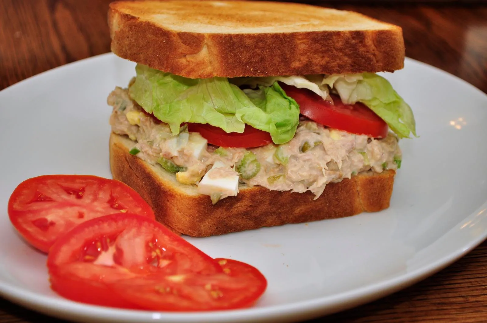 Close-up of a freshly made tuna sandwich with lettuce and tomato on toasted whole grain bread, served on a wooden board.