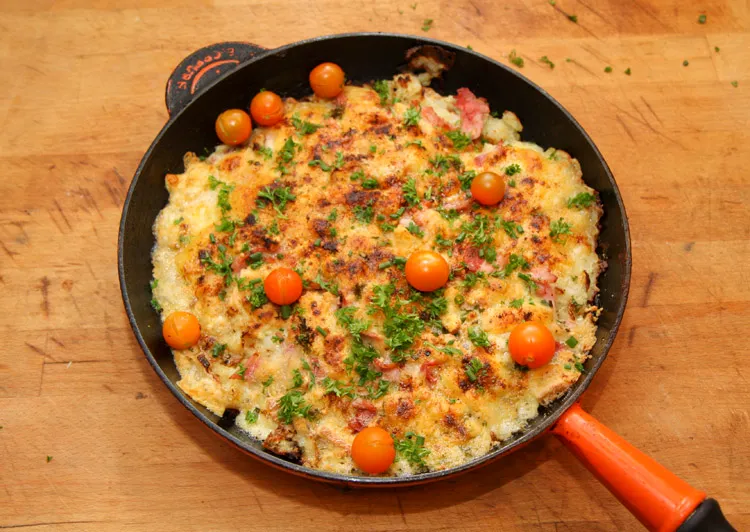 Cast iron skillet filled with crispy bacon, golden breakfast potatoes, sunny-side-up eggs, and melted cheddar cheese, garnished with fresh chives.