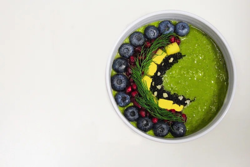 Green protein smoothie bowl with banana, avocado, spinach, and colorful berry toppings in a white bowl.