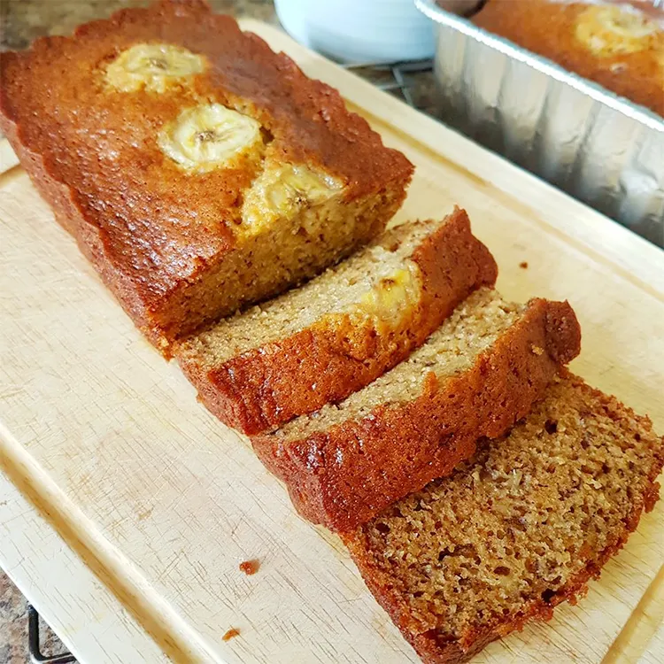 Slice of moist banana bread on a wooden cutting board with ripe bananas beside it