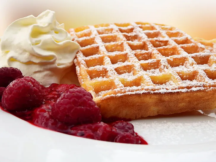 Close-up of a golden Belgian waffle topped with fresh strawberries, blueberries, whipped cream, and maple syrup.
